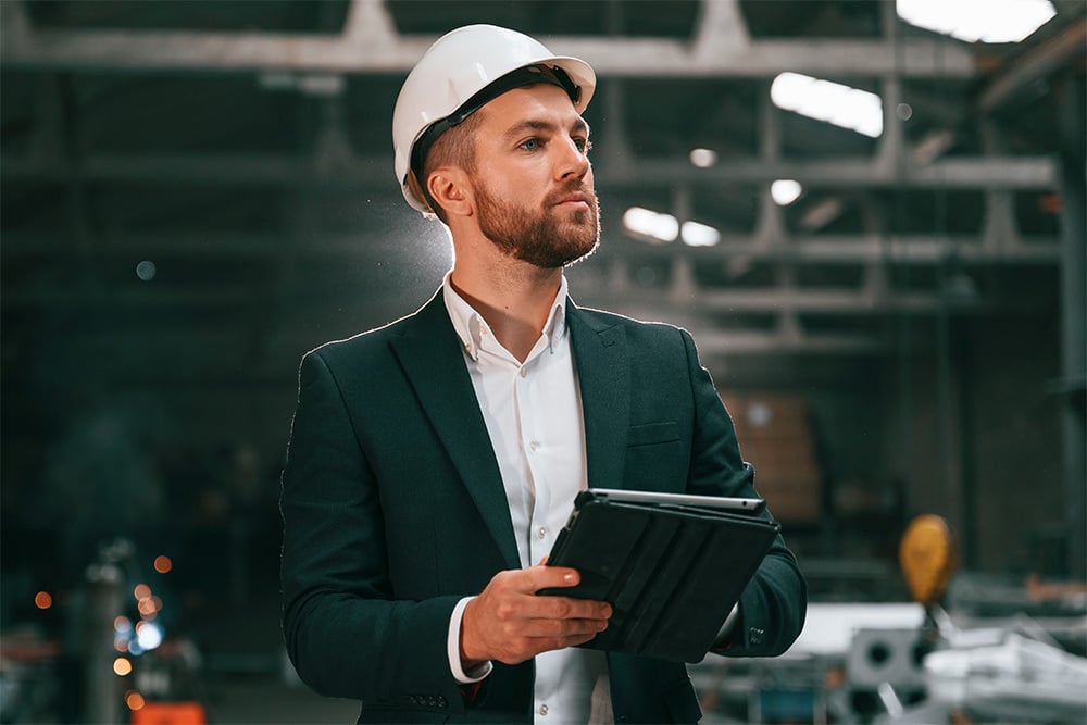 Unternehmer im Anzug mit Schutzhelm, ein Tablet in der Hand, in einer Werkshalle.